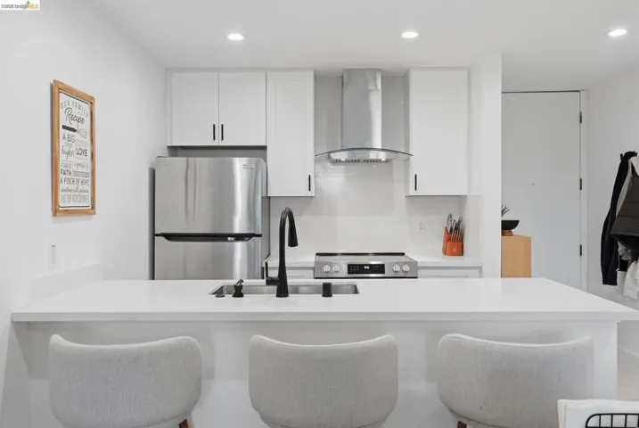 Contemporary kitchen featuring white cabinetry, stainless steel appliances, a counter-depth refrigerator, built-in range with overhead ventilation, and a spacious island with a sink and matte black faucet