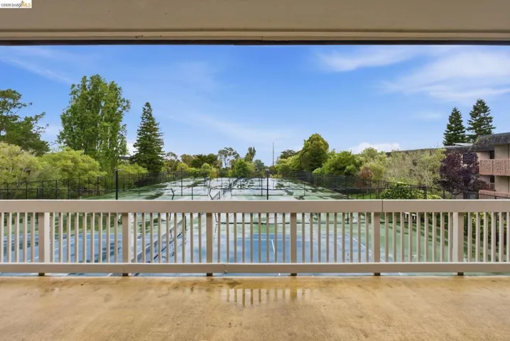Balcony with concrete flooring and a white railing overlooking multiple tennis courts, surrounded by mature trees and lush landscaping