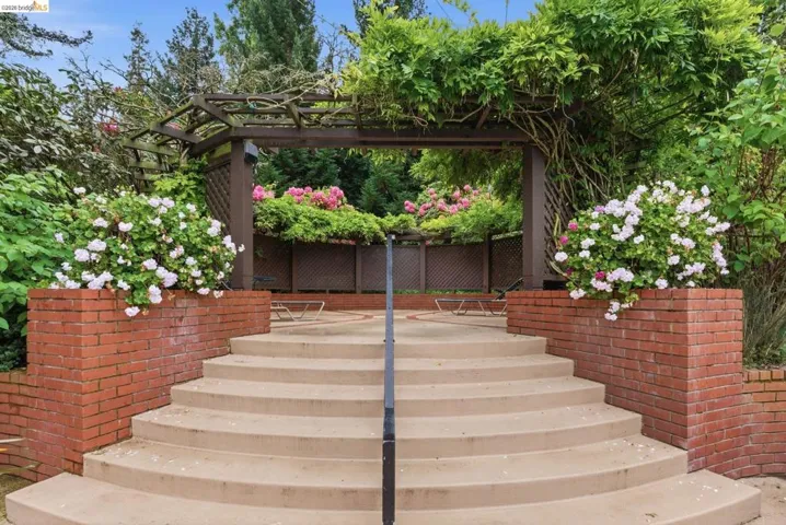 Curved concrete steps ascend to a pergola draped with mature greenery, featuring integrated brick planters with flowering shrubs