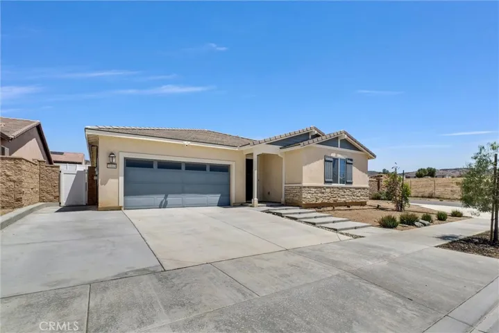 The main photo features an attractive single-story home with clean curb appeal and a modern exterior design. The property showcases a wide concrete driveway leading to a two-car garage with decorative window panels, complemented by stone accents along the front façade. A covered entryway adds character, while drought-tolerant landscaping and a spacious front yard provide a low-maintenance touch. The home sits in a peaceful setting with open views and a bright blue sky creating a welcoming first impression.