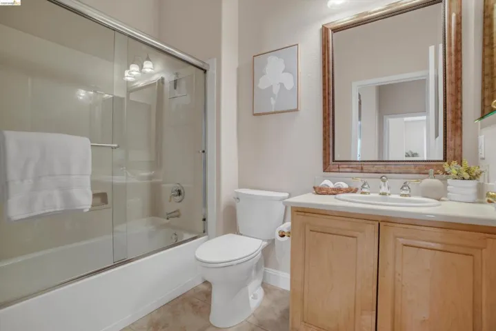 Bathroom featuring a glass-enclosed shower-over-tub, a single vanity with a light-toned countertop, a toilet, and tile flooring