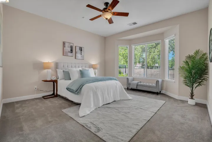 Spacious bedroom featuring a bay window, ceiling fan with integrated lighting, neutral wall tones, and plush carpeting