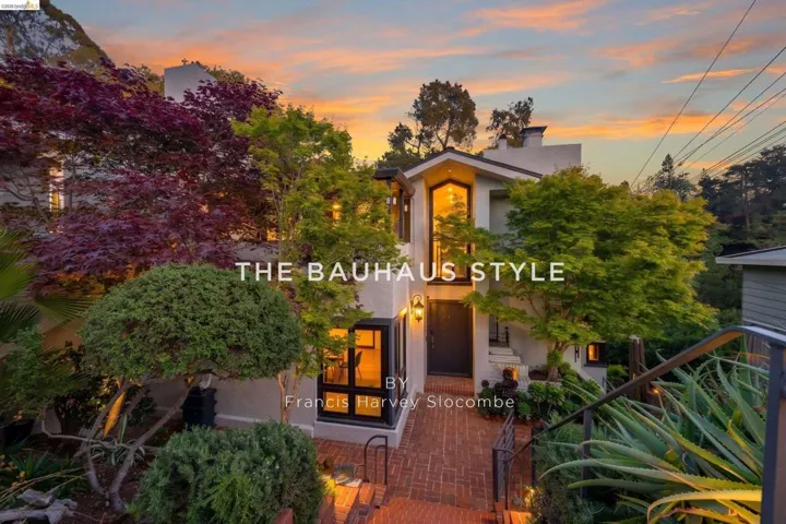 Multi-level residence featuring a brick pathway, mature landscaping, exterior lighting, an arched upper window, and a dark entry door