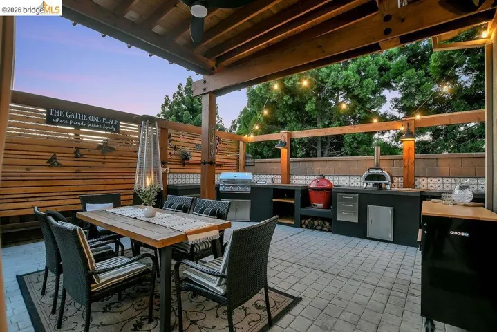 Expansive patio featuring a built-in outdoor kitchen with integrated appliances, a wood-slat privacy fence, overhead string lighting, and a wood pergola with a ceiling fan