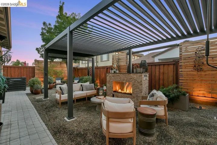 Spacious outdoor living area featuring a modern black pergola, stone-clad fireplace, gravel ground cover, and a paved walkway