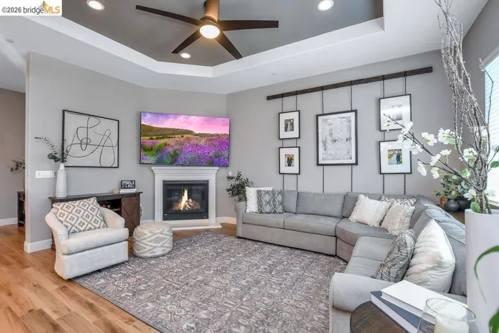 Living room featuring a tray ceiling with recessed lighting and a ceiling fan, a fireplace with a white mantel, and wood-finish flooring