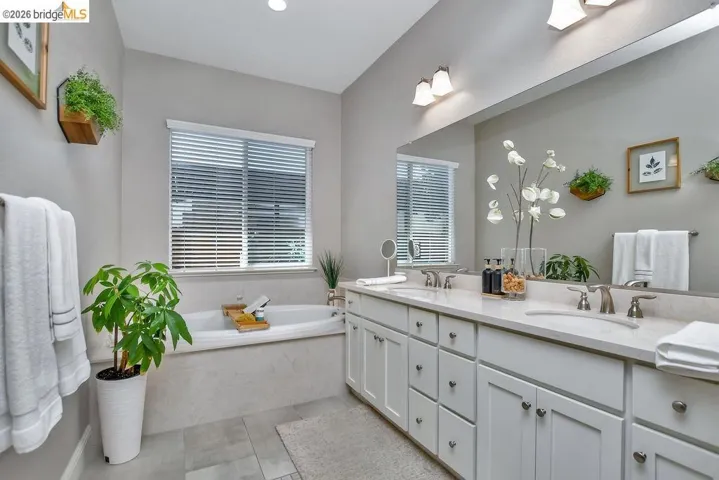 Spacious bathroom featuring a built-in soaking tub with a surrounding tile deck, dual sink vanity with white cabinetry, and an expansive wall-mounted mirror
