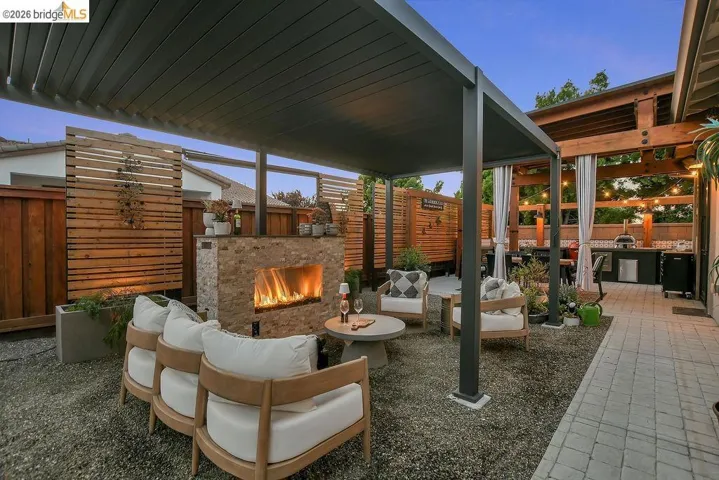 Outdoor living space featuring a louvered pergola, stone-clad fireplace, gravel patio area, wood privacy screens, and a built-in outdoor kitchen with a paver pathway