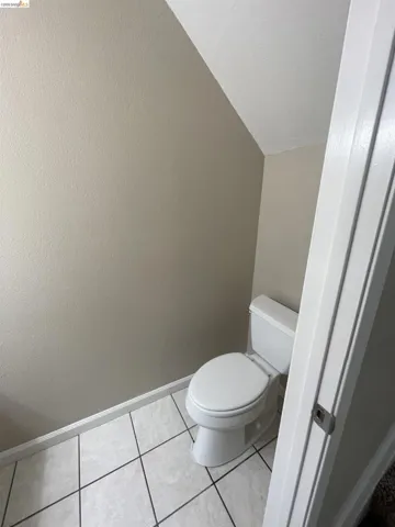 White toilet featuring a standard-height bowl, light beige textured walls, white baseboards, and light-colored square tile flooring with dark grout