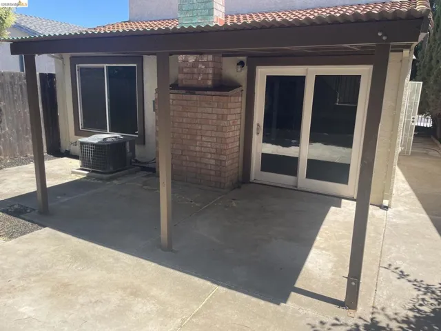Outdoor patio area featuring a covered structure with a corrugated roof, a brick chimney, a large sliding glass door, an additional window, and a concrete slab surface