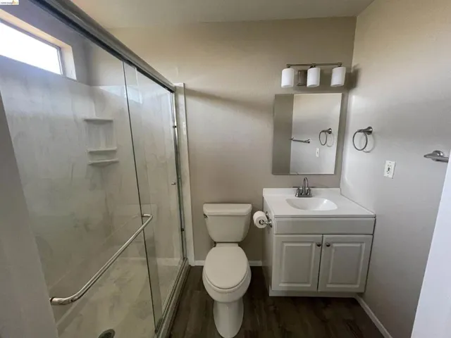 Bathroom featuring a glass-enclosed shower with integrated shelving, a white single-sink vanity with cabinet storage, a wall-mounted mirror, and wood-finish flooring