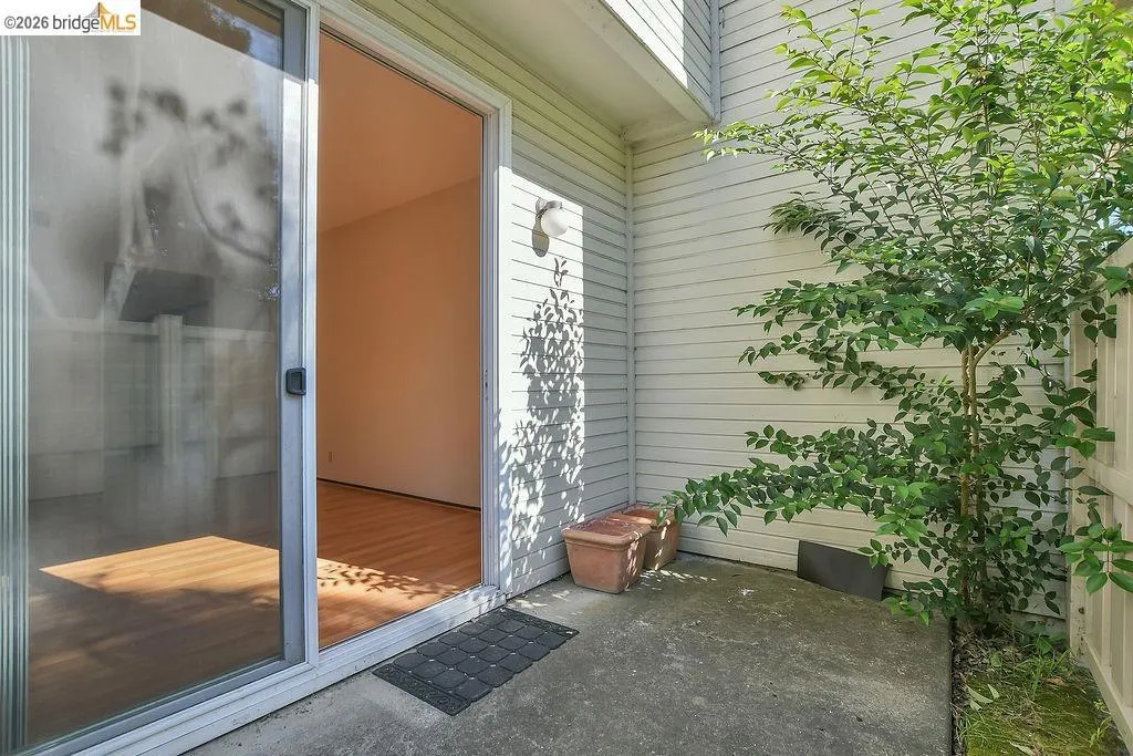 Sliding glass door opening to a concrete patio area, featuring a mature shrub, white horizontal siding, and an outdoor wall sconce