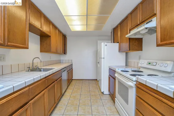 Galley kitchen featuring wood cabinetry, white tile countertops, and a full suite of white appliances