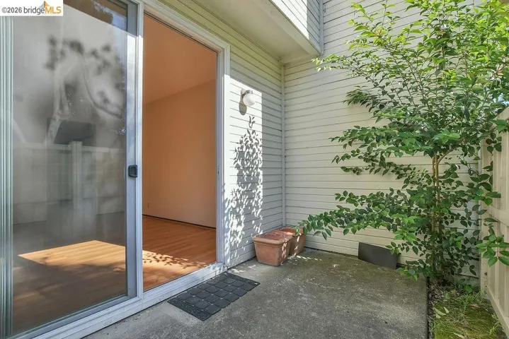 Sliding glass door opening to a concrete patio area, featuring a mature shrub, white horizontal siding, and an outdoor wall sconce