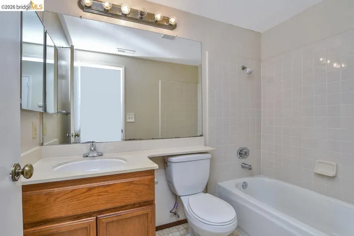 Bathroom featuring a light wood-finish vanity with an integrated sink, a large wall-mounted mirror, overhead vanity lighting, a white toilet, and a bathtub with tiled surround