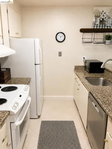 Compact kitchen featuring granite countertops, a stainless steel sink, tile flooring, white cabinetry, and a built-in dishwasher