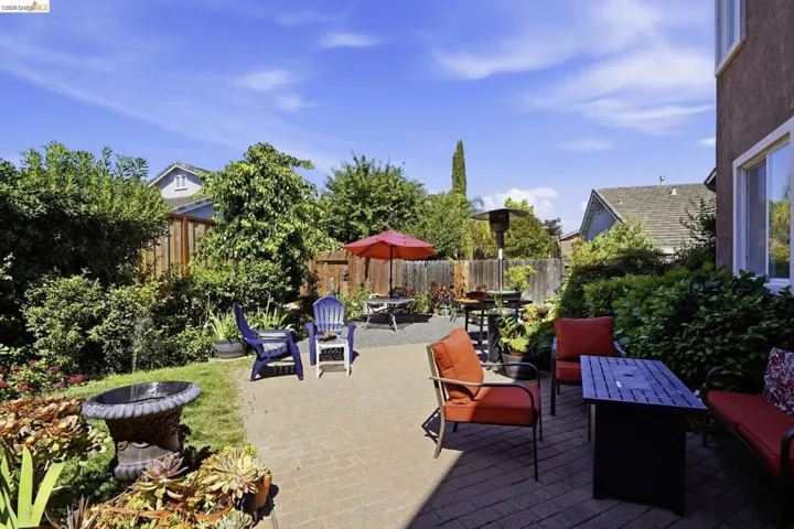 Expansive backyard featuring a brick paver patio, a gravel seating area with a red umbrella, established green landscaping, and a stucco exterior with multiple windows