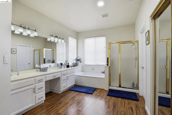 Bathroom featuring wood-finish flooring, a double vanity with ample counter space, a built-in soaking tub, and a shower with a brass-finish frame