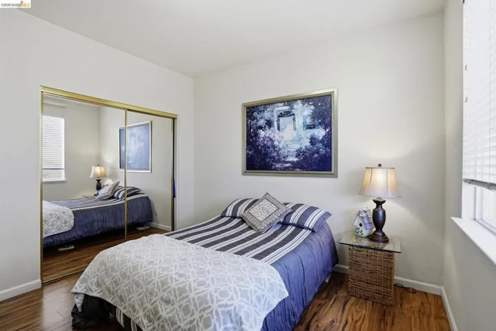 Bedroom featuring wood-finish flooring, a window with horizontal blinds, and a built-in mirrored closet with gold-toned trim