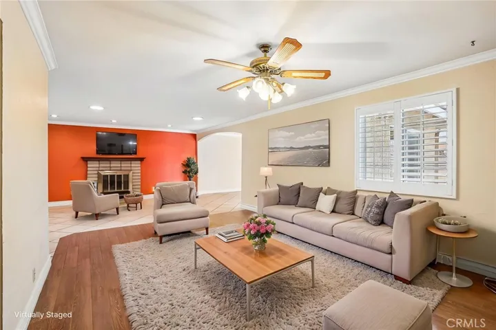 Virtual staged Living Room - This spacious living room offers an open and versatile layout with warm wood-style flooring, recessed lighting, and abundant natural light throughout. A cozy brick fireplace creates a welcoming focal point, complemented by a bold accent wall that adds character and charm. Plantation shutters, crown molding, and the seamless flow into adjacent living spaces make this room ideal for both relaxing and entertaining