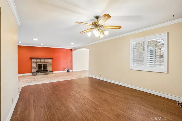 Living Room - This spacious living room offers an open and versatile layout with warm wood-style flooring, recessed lighting, and abundant natural light throughout. A cozy brick fireplace creates a welcoming focal point, complemented by a bold accent wall that adds character and charm. Plantation shutters, crown molding, and the seamless flow into adjacent living spaces make this room ideal for both relaxing and entertaining