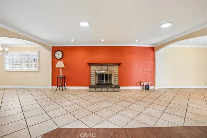 Living Room - This spacious living room offers an open and versatile layout with warm wood-style flooring, recessed lighting, and abundant natural light throughout. A cozy brick fireplace creates a welcoming focal point, complemented by a bold accent wall that adds character and charm. Plantation shutters, crown molding, and the seamless flow into adjacent living spaces make this room ideal for both relaxing and entertaining