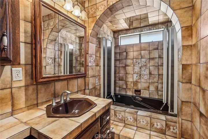Bathroom 1 - This uniquely designed bathroom showcases vintage charm and custom craftsmanship with floor-to-ceiling decorative tile work and an eye-catching arched shower enclosure. Rich wood cabinetry and warm earth-tone finishes create a cozy, rustic ambiance, while the tiled vanity and detailed floral accents add character throughout the space. The spacious shower/tub combination, natural light, and abundant tile surfaces provide both functionality and timeless personality.