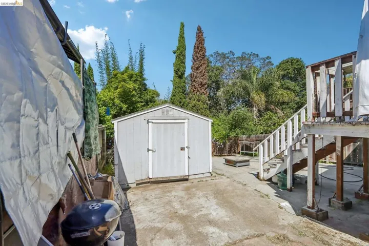 Paved outdoor area featuring a storage shed, wood deck structure with stairs, and mature trees