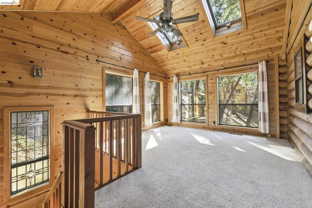 Bedroom /loft with carpet flooring, knotty pine walls, vaulted ceiling, ceiling fan, and large windows. Open railing detail visible from the loft connection.
