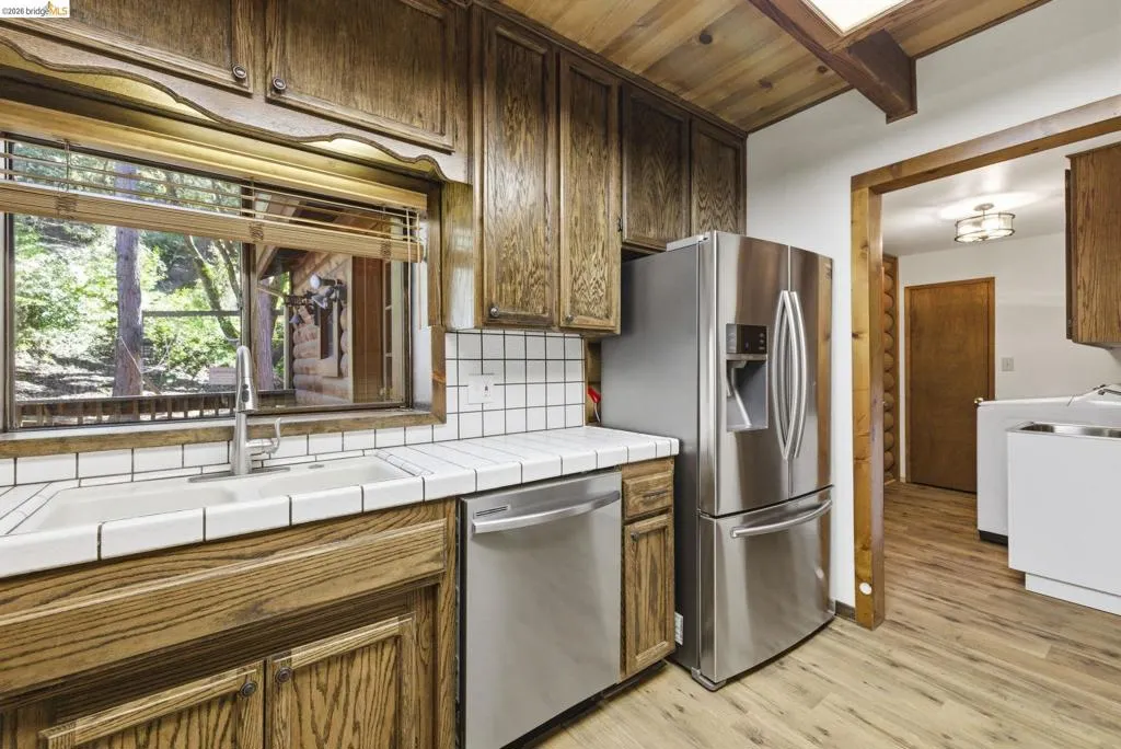 Kitchen showing stainless steel refrigerator, dishwasher, and tile countertops. Natural light from window above sink, with wood cabinetry and open access to main living area.