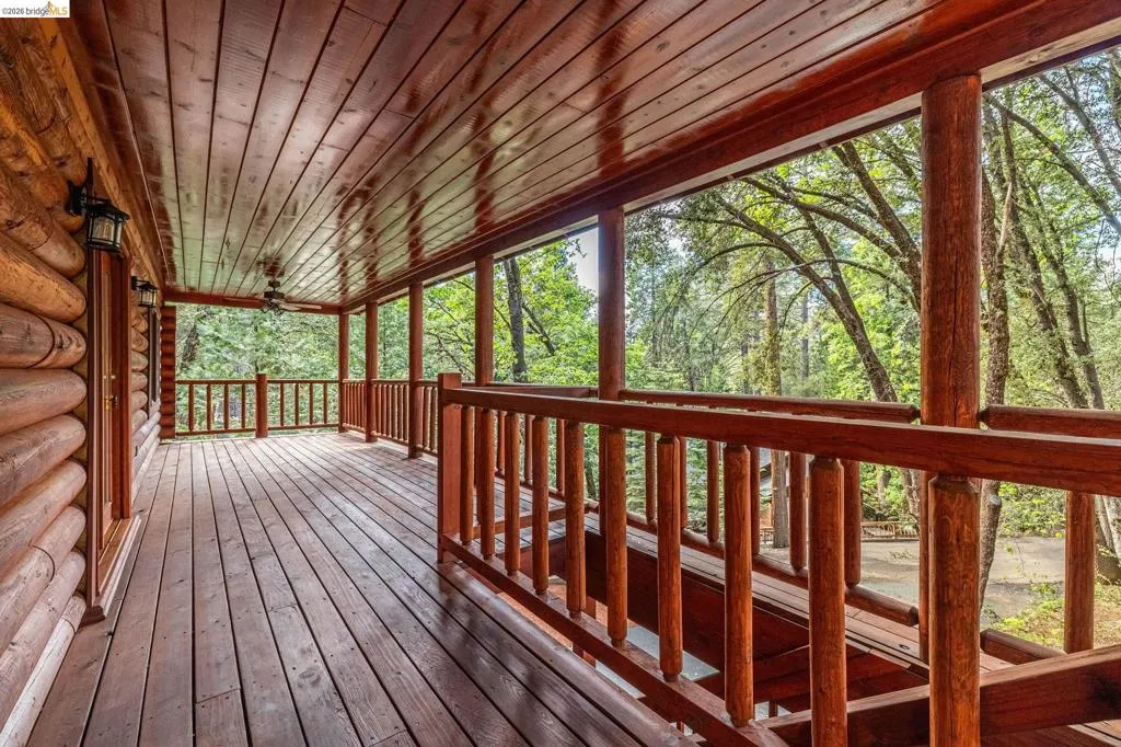 Covered wrap-around deck with wood plank flooring and wood railing on three sides. Open views into the surrounding pine canopy with natural light throughout.