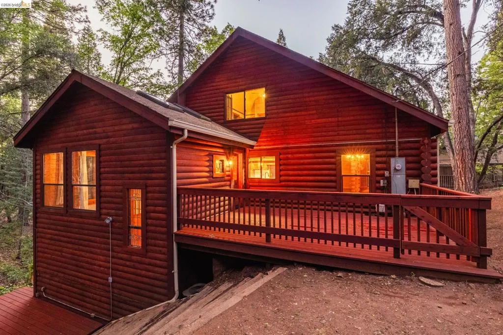 Side-angle twilight shot showing deck depth, wood railing, and lower level access. Mature pines and natural surroundings frame the property at dusk.