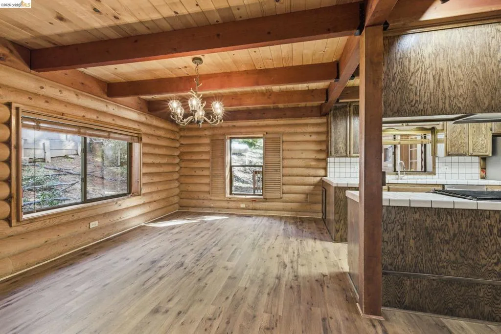 Main living area with decorative chandelier, exposed ceiling beams, knotty pine walls, and open flow to kitchen. Large windows along the back wall.