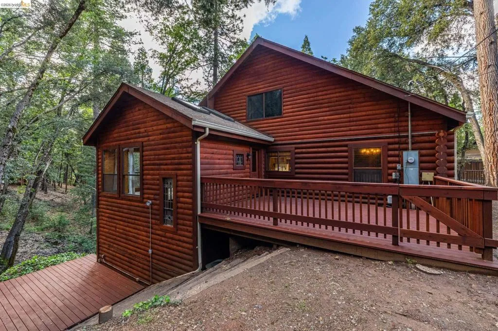 Rear exterior view showing multi-level structure, lower deck, and attached garage bay. Wooded lot with mature pines and driveway access visible.