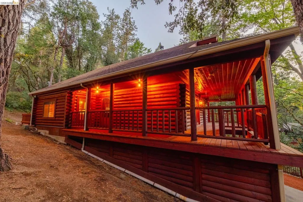Wide twilight exterior showing full structure against the pine tree backdrop. Warm interior light visible through multiple windows across all three levels.