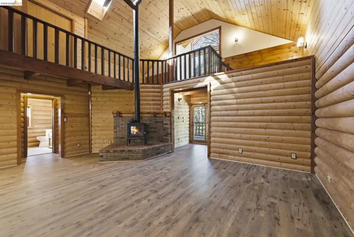 Open great room with vaulted ceiling, exposed wood beam, knotty pine walls, wood-look flooring, and second-floor loft railing above. Wood-burning fireplace visible at left. Large windows bring in natural light.