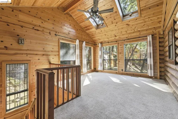 Bedroom /loft with carpet flooring, knotty pine walls, vaulted ceiling, ceiling fan, and large windows. Open railing detail visible from the loft connection.