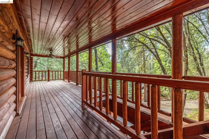 Covered wrap-around deck with wood plank flooring and wood railing on three sides. Open views into the surrounding pine canopy with natural light throughout.