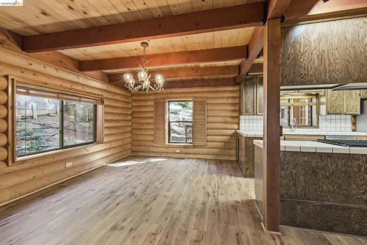 Main living area with decorative chandelier, exposed ceiling beams, knotty pine walls, and open flow to kitchen. Large windows along the back wall.