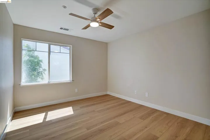 Spacious room featuring wood-finish flooring, a large window, neutral wall tones, white trim, and a ceiling fan with integrated lighting
