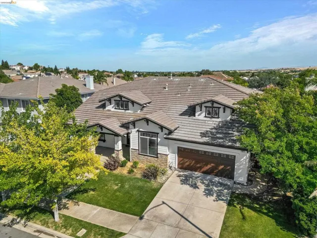 Contemporary residence featuring a brown tile roof, dormer windows, a 3 tandem car garage with a wood-finish door, stone veneer accents, and mature landscaping