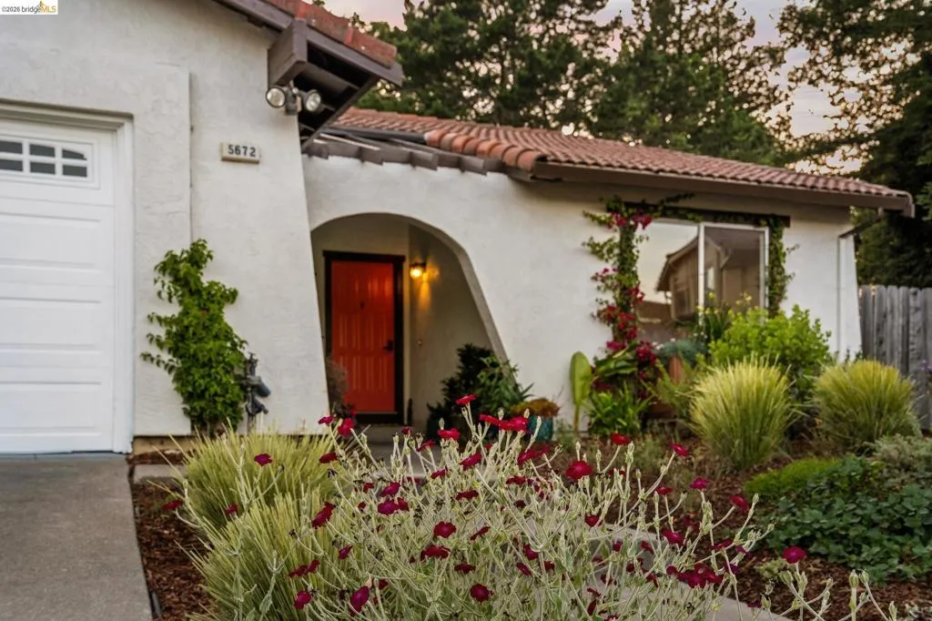Stucco exterior featuring a terracotta tile roof, prominent arched entryway with an orange door, a white garage door, and mature landscaping with varied plantings