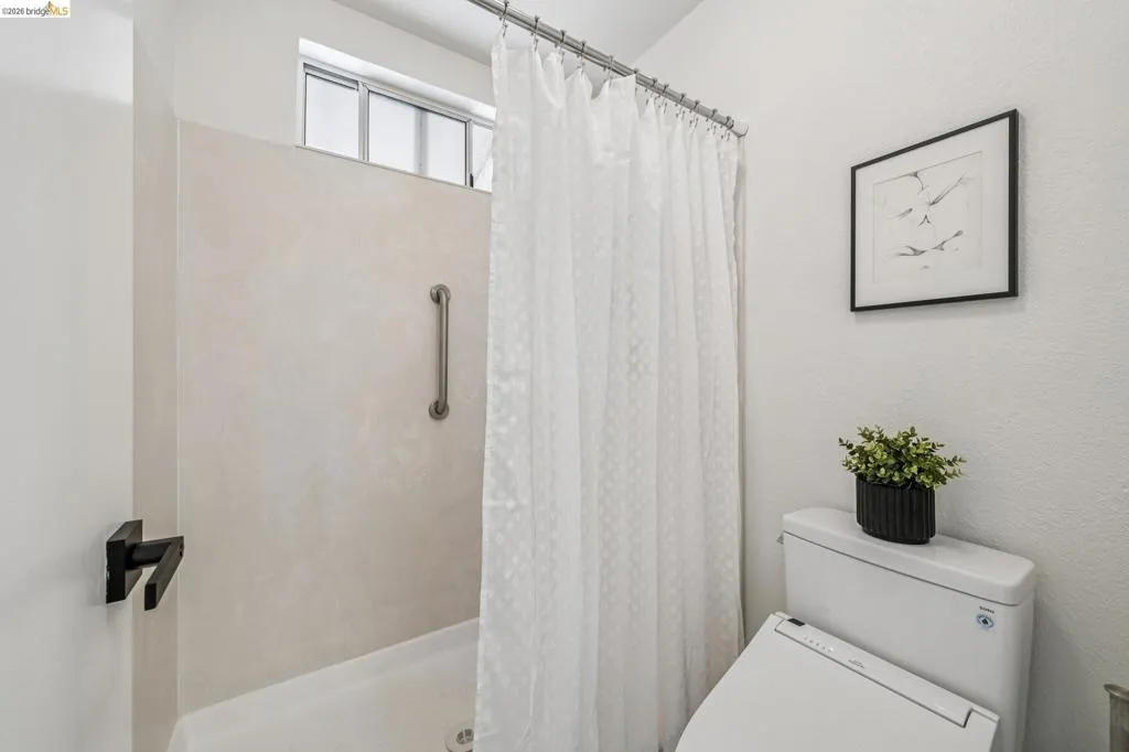 Shower stall with a grab bar, featuring a transom window, white shower curtain, and a white toilet