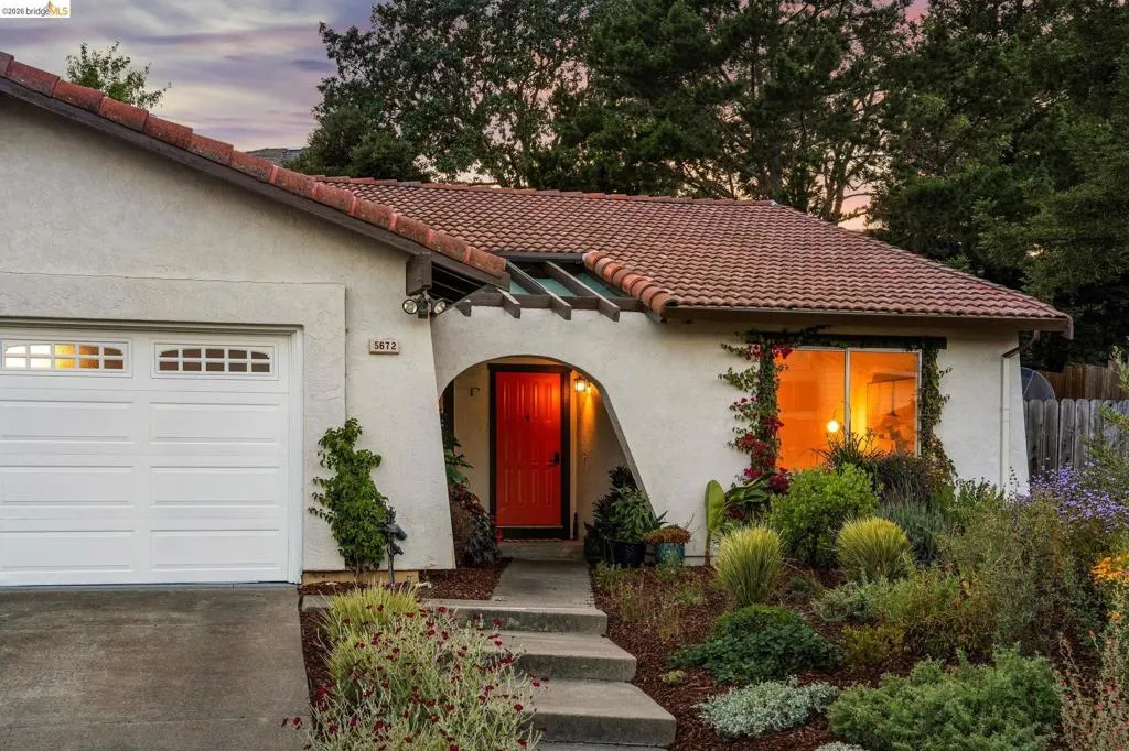 Stucco exterior with a terracotta tile roof, featuring a white garage door with upper window inserts, an arched entryway with a vibrant red door, and extensive front landscaping with mature plantings