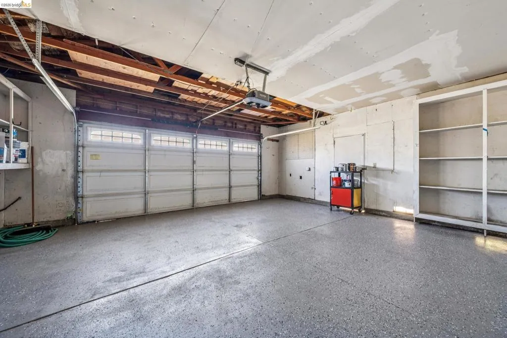 Spacious garage featuring a durable epoxy-coated floor, automatic garage door with window inserts, exposed beam ceiling, and built-in shelving for organization