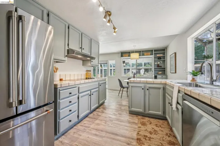 Kitchen featuring light gray cabinetry, stainless steel appliances, wood-finish flooring, tiled countertops, and a built-in shelving unit