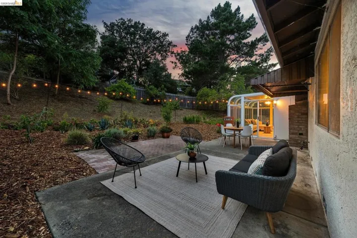 Outdoor patio featuring a glass-enclosed sunroom, concrete patio, string lighting, and terraced landscaping with mature plantings