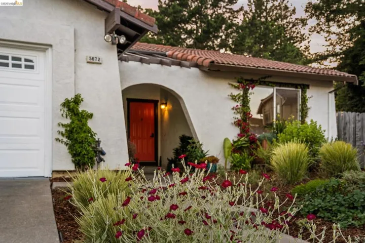 Stucco exterior featuring a terracotta tile roof, prominent arched entryway with an orange door, a white garage door, and mature landscaping with varied plantings