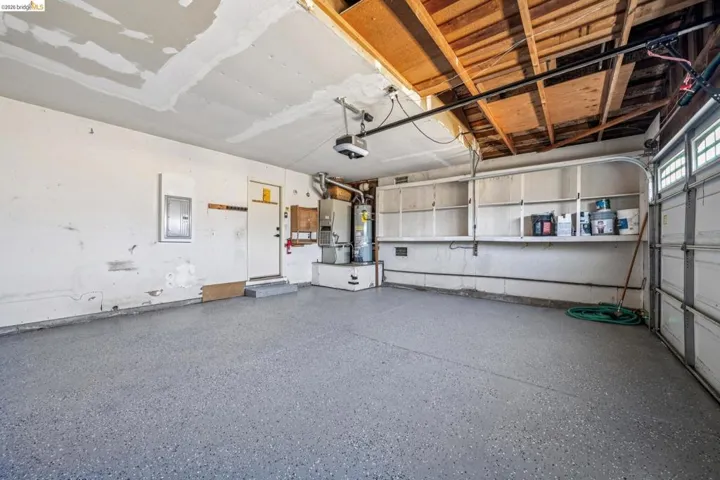 Garage featuring an epoxy-coated concrete floor, exposed wood ceiling beams, built-in shelving, an automatic garage door opener, and a water heater installation