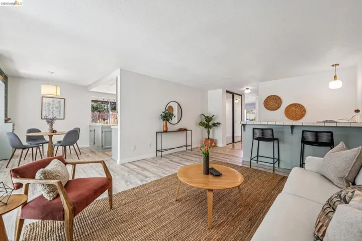 Open-concept living space with light-tone wood-finish flooring, a breakfast bar featuring a tile countertop, a dining area with a contemporary chandelier, and a kitchen with light-colored cabinetry visible through a pass-through